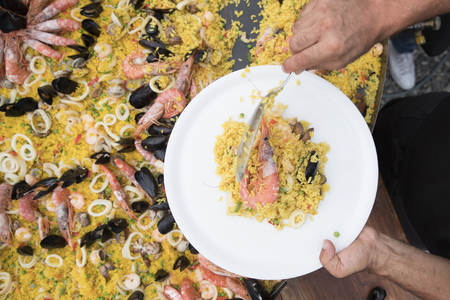top view close up of a man serving paella with a spoon in his hand next to a big paelleraの写真素材