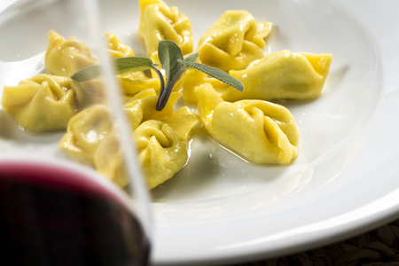a plate of tortelli, a typical stuffed pasta from Piacenza, north Italy, with butter and fresh sage leafs. the plate is hidden by a glass of red wineの写真素材