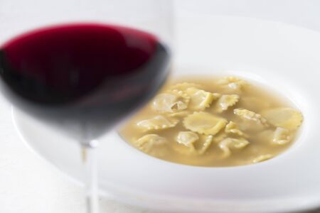a plate of Anolini, a typical filled pasta of Piacenza, north italy,  served in hot beef broth. the plate stands behind a glass of red wineの写真素材