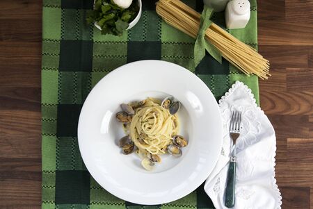 flat horizontal shot of a plate of spaghetti with clams on a green table sheet with raw spaghetti on topの写真素材