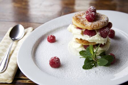 horizontal shot of a creamy and fresh dessert made with raspberry, puff pastry layers and fluffy cream. the plate is on a dark wooden table with a spoon on the leftの写真素材