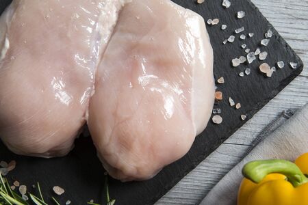 top view  closeup of a raw italian chicken breast on a black slate chop board with rosemary and a raw yellow and red  pepper and pink himalaya salt grain.の写真素材