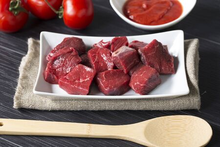frontal shot of a tray with big pieces of raw beaf with tomatoes and a wooden spoon.の写真素材
