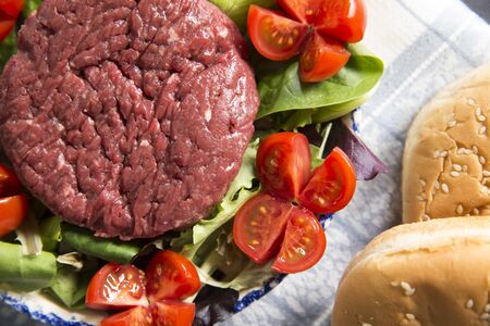 top view closeup of a plate with a raw hamburger on it on a lettuce and tomatoes salad with hamburger breads.の写真素材