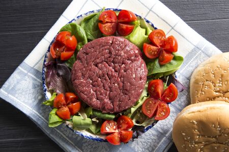 top view shot of a plate with a raw hamburger on it on a lettuce and tomatoes salad with hamburger breads.の写真素材