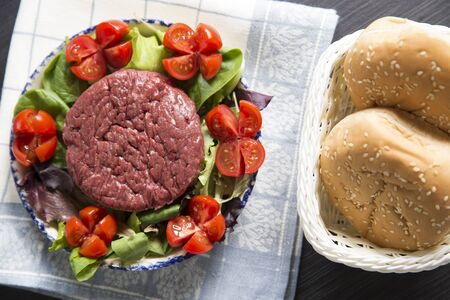 top view shot of a plate with a raw hamburger on it on a lettuce and tomatoes salad with hamburger breads on the right.の写真素材