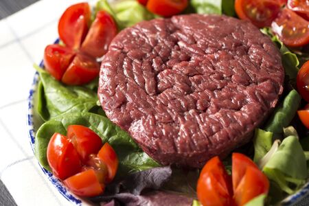 horizontal closeup of a plate with a raw hamburger on it on a lettuce and tomatoes salad.の写真素材