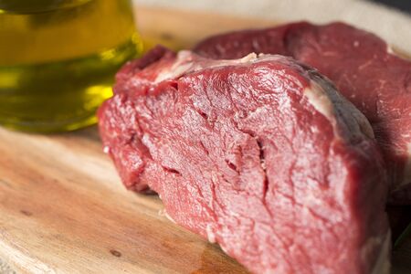 horizontal closeup of a wooden chop board with two slices of raw beef on it and a bottle of olive oil on a dark wooden table.の写真素材