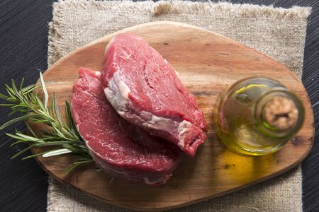 top view shot of a wooden chop board with two slices of raw beef on it and a bottle of olive oil on a dark wooden table.の写真素材