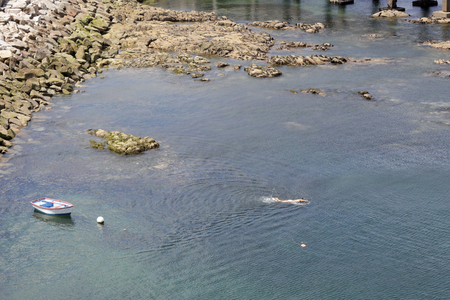 A small white wooden boat anchored in a port next to a rock pier and cliffs with some buoys and a swimming manの写真素材