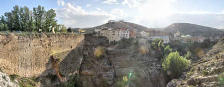 A landscape at the sunset of the rural country town of Almonacid de la Cuba, in Spain, with houses on a small rocky valley with the Rio Aguavivas river and waterfall at its bottom. An ancient Roman bridge is crossing the river.の写真素材