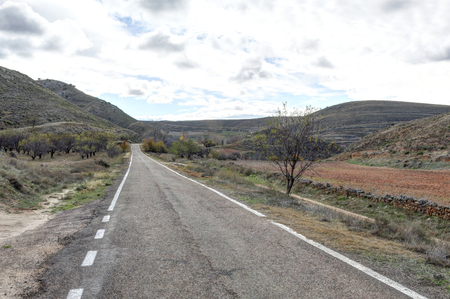A paved two ways street going into the Aragonese countryside hills, with bare trees and green fields, in a sunny autumn, around Monterde town, Spainの写真素材
