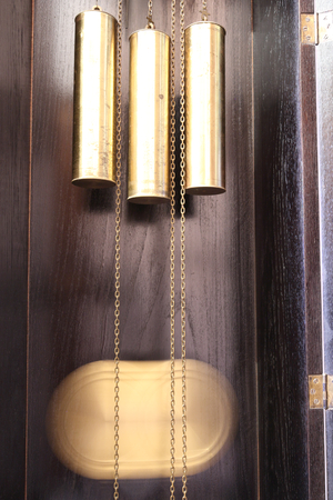 The motion blur of a golden round pendulum clock weight moving inside a black wooden box with three yellow metal weights hanging on chain cordsの写真素材
