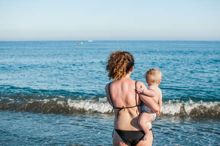 mother and baby with their backs on the beach. Woman with curly hair and blond boy looking out to...