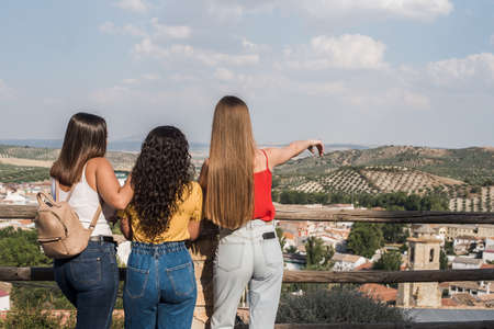 Three happy young tourist friends back to back looking at a village landscapeの写真素材