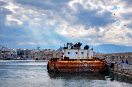 Heraklion, Crete - Greece. Floating Crane at the port of Heraklion. Fortress Koules and part of Heraklion at the background. Sun rays breaking through the clouds of a dramatic sky over the cityのeditorial素材