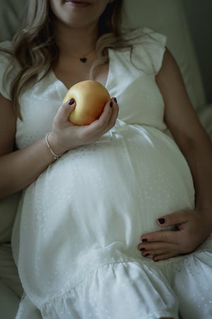 Pregnant white woman in a white dress on the couch eating an appleの写真素材