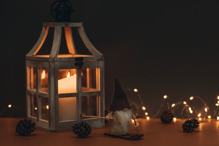A cute dwarf, in a high hat, on skis and on a brown background, decorated with lanterns with a candle, cones and a garland. Suitable for postcards and Christmas greetingsの写真素材