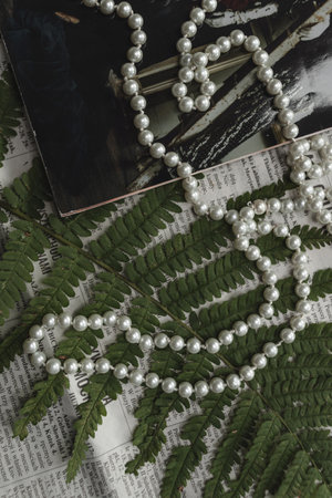 A string of pearls lies against the background of a green fern leaf, a magazine and a newspaper.