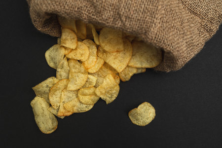 A canvas bag with potato chips scattered across a black table. Potato chips spilled out of bagの写真素材