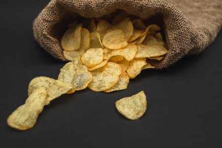 A canvas bag with potato chips scattered across a black table. Potato chips spilled out of bagの写真素材