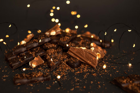 <p>Pieces of dark and milk chocolate lie in a pile on a dark paper background. In the background,...