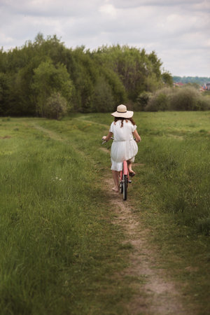 A girl in a white dress and a hat rides a bicycle along a path in a fieldの写真素材