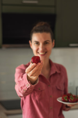 A sweet smiling mature woman holds out a strawberry. Strawberry in the hands of a womanの写真素材