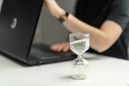 Hourglass in the foreground. The woman opens the laptop to start working. The beginning of the working day.の写真素材