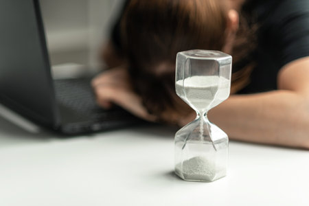 Hourglass in the foreground. A woman is sitting in front of a laptop and is stressed, covering her face with her hands. The concept of lack of time. Deadline not metの写真素材