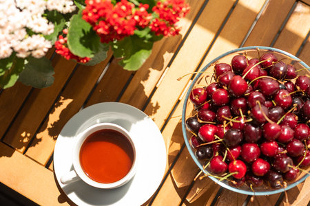Cherries in a large glass bowl on a wooden table. Next to a cup of tea and kalanchoe flowers in a white planter. Sunny summer day on the balconyの写真素材