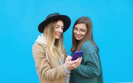 Girls in a blue background taking selfie - Teenagers posing cheerfully with mobile phone in hand -...