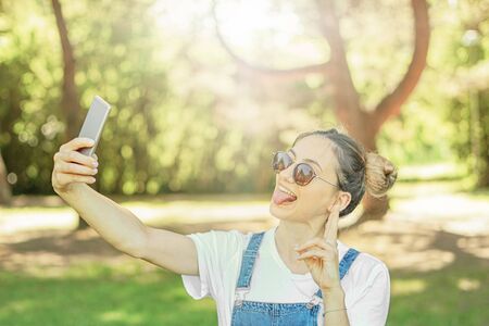 Happy cheerful young girl in nature taking selfie with space bun hairstyle - Teeneger doing funny face and holding her smartphoneの写真素材
