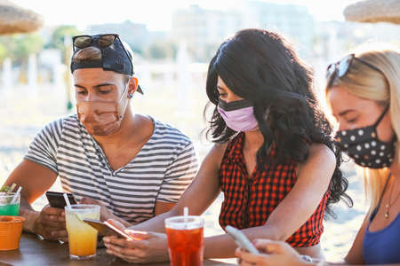 Friends using smartphone in beach bar with face mask on - Young people using mobile device in...
