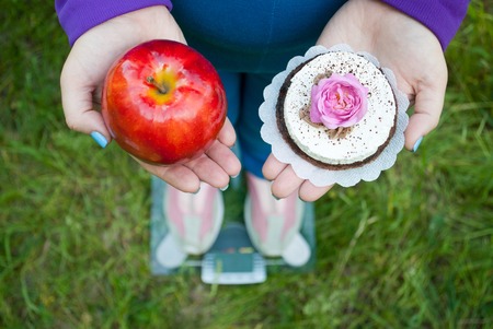 Fat woman wants to lose weight diet top view in blue suit stands on a transparent glass scales in pink sneakers on a green grass selects red big brown or brown brown with white cake with rose of pink color holding them in hands with short blue nails on blの写真素材