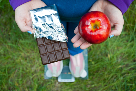 Fat woman wants to lose weight diet top view in blue suit stands on transparent glass scales in pink sneakers on green grass selects big red apple or chocolate bar in foil chocolatesの写真素材