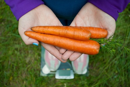 Fat woman wants to lose weight diet top view in blue suit stands on transparent glass scales in pink sneakers on green grass holds in folded folds three large orange fresh carrots with short blue nails on blused backgroundの写真素材