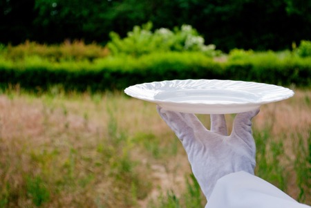 A waiter's hand in a white glove and with a white napkin holding an empty glass white big plate on a...