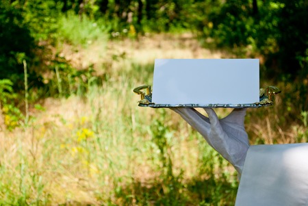 A waiter's hand in a white glove and with a white napkin holding a metal tray of silvery rectangular metal with a white sign on a blurred background of nature green bushes and treesの写真素材