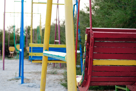 Multicolored childrens swing from metal, high, against a background of green tree bush, on sand summer sun, playground, blue skyの写真素材