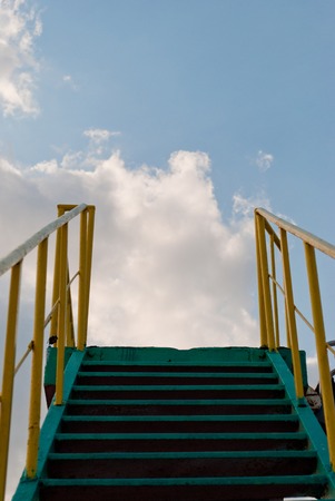 A green ladder of metal leads upwards, against a blue sky, white clouds, a yellow metal railing