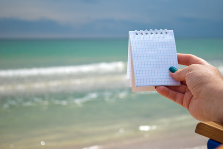 female hand girl with manicure holds notepad for notes in a cage on a sea background beach waves...