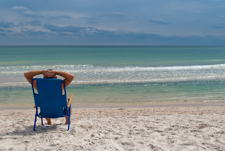 young man in a straw hat sits on the seashore from the back, summer beach waves sky with clouds, sand, sits in a deckchair throwing his hands behind his head and looking at the horizon, resting on a summer holiday, vacationの写真素材