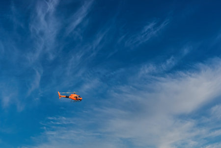 A helicopter flying against a blue sky with white clouds, showing motion and aircraft detail in...