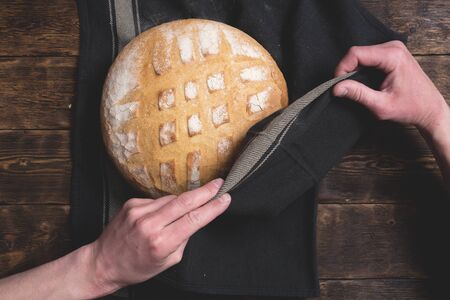 Baker is holding in hands a fresh baked bread on a wooden table background.の写真素材