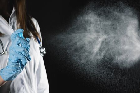 Woman doctor is spraying a antiseptic from a bottle in her hands on dark background.