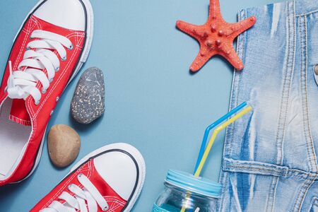 Red gum shoes, jeans shorts and cocktail jar with tube on blue flat lay background. Summer vacation...