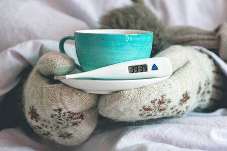Flu sick woman with thermometer and in mittens warms up with hot tea with lemon.