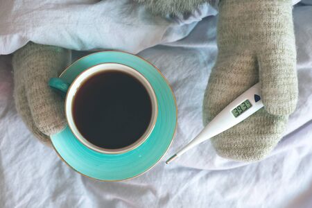 Flu sick woman with thermometer and in mittens warms up with hot tea with lemon.の写真素材