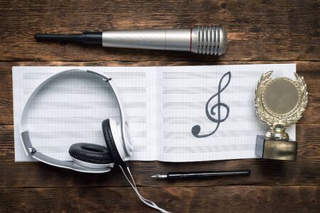 Music award concept background. Musical book with a treble clef, golden medal award trophy, microphone and headphones on wooden table.の写真素材